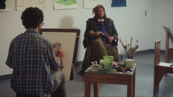 Movie still from “Showing Up” (2022), directed by Kelly Reichardt – An older woman sitting in front of an easel in front of an older man; Medium shot, Over the shoulder angle