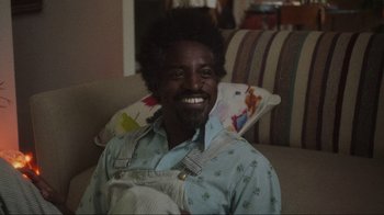 Movie still from “Showing Up” (2022), directed by Kelly Reichardt – A man sitting on a couch smiling for the camera; Close Up shot, High angle