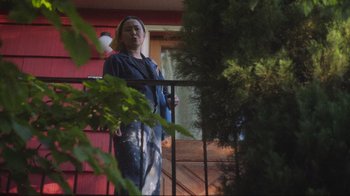 Movie still from “Showing Up” (2022), directed by Kelly Reichardt – A woman standing on a porch holding a broom; Wide shot, Low angle