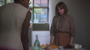 Movie still from “Showing Up” (2022), directed by Kelly Reichardt – A woman standing next to a table with food on top of it; Medium shot, Over the shoulder angle
