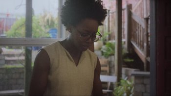 Movie still from “Showing Up” (2022), directed by Kelly Reichardt – A woman with glasses is looking down at a plant; Medium shot, Over the shoulder angle