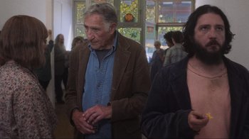 Movie still from “Showing Up” (2022), directed by Kelly Reichardt – An older man standing next to a group of people in a room; Medium shot, Over the shoulder angle