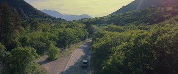 Movie still from “Sick” (2022), directed by John Hyams – A car driving down a road surrounded by green trees; Extreme Wide shot, High angle
