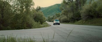 Movie still from “Sick” (2022), directed by John Hyams – A car driving down a road near some trees; Extreme Wide shot, High angle