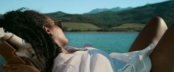 Movie still from “Sick” (2022), directed by John Hyams – A woman with sunglasses is laying on the ground by the water; Medium shot, High angle