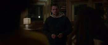 Movie still from “Sick” (2022), directed by John Hyams – A man standing in a room holding a cell phone; Medium shot, Over the shoulder angle