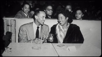 Movie still from “Sidney” (2022), directed by Reginald Hudlin – A man and a woman sitting at a table with drinks; Medium shot, High angle