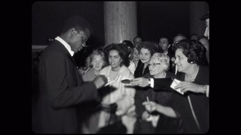 Movie still from “Sidney” (2022), directed by Reginald Hudlin – A black and white photo of a man signing autographs for a group of people; Medium shot, Over the shoulder angle
