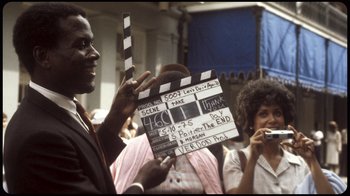 Movie still from “Sidney” (2022), directed by Reginald Hudlin – A man holding a clapperboard in front of a crowd of onlookers; Close Up shot, Over the shoulder angle