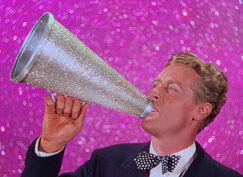 Movie still from “Singin' in the Rain” (1952), directed by Gene Kelly – A man in a suit and bow tie is holding up a large silver speaker; Close Up shot, Low angle