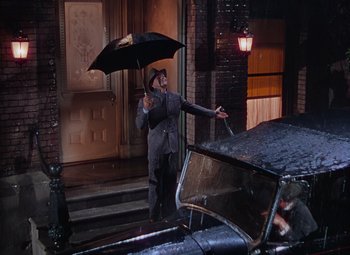 Movie still from “Singin' in the Rain” (1952), directed by Gene Kelly – A man holding an umbrella while standing next to an old car; Wide shot, High angle
