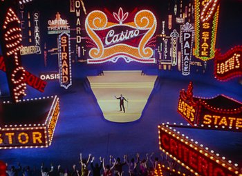 Movie still from “Singin' in the Rain” (1952), directed by Gene Kelly – A man standing on a stage in front of neon signs; Extreme Wide shot, High angle