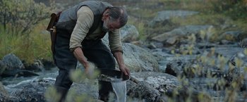 Movie still from “Sisu” (2022), directed by Jalmari Helander – An old man in a brown vest and black pants is digging a hole in the rocks; Medium shot, High angle