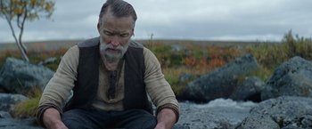 Movie still from “Sisu” (2022), directed by Jalmari Helander – An older man sitting on the ground near some rocks; Medium shot, Over the shoulder angle