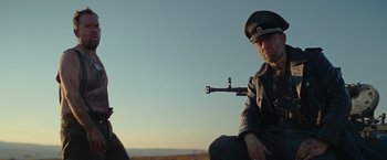 Movie still from “Sisu” (2022), directed by Jalmari Helander – A man sitting on top of a hill holding a rifle; Medium shot, Low angle