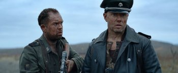 Movie still from “Sisu” (2022), directed by Jalmari Helander – A man in a military uniform holding a gun next to another man in a military uniform; Medium shot, Over the shoulder angle