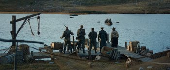 Movie still from “Sisu” (2022), directed by Jalmari Helander – A group of men standing next to a body of water; Wide shot, High angle