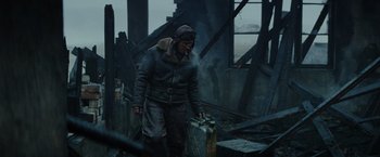 Movie still from “Sisu” (2022), directed by Jalmari Helander – A man in a jacket and hat holding a suitcase in the middle of a burned area; Medium shot, Low angle