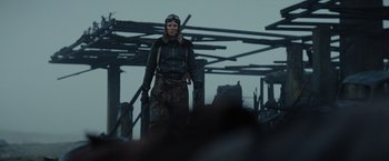 Movie still from “Sisu” (2022), directed by Jalmari Helander – A man in a leather jacket standing in front of a building; Medium shot, Low angle