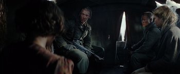 Movie still from “Sisu” (2022), directed by Jalmari Helander – A man sitting in a dark room holding a rifle; Medium shot, Over the shoulder angle