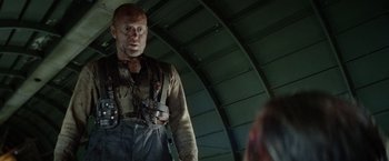 Movie still from “Sisu” (2022), directed by Jalmari Helander – A bald man in a brown shirt is standing in an airplane hangar; Medium shot, Low angle