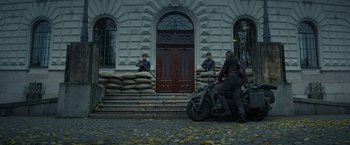Movie still from “Sisu” (2022), directed by Jalmari Helander – A man riding a motorcycle in front of a building; Wide shot, Low angle