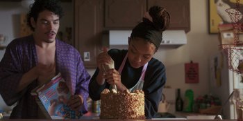 Movie still from “Sitting in Bars with Cake” (2023), directed by Trish Sie – A young woman is decorating a cake in the kitchen; Medium shot, Over the shoulder angle