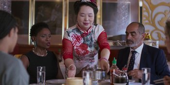 Movie still from “Sitting in Bars with Cake” (2023), directed by Trish Sie – An asian woman cutting a cake at a table; Medium shot, Over the shoulder angle