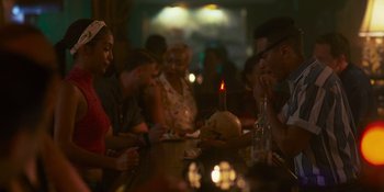 Movie still from “Sitting in Bars with Cake” (2023), directed by Trish Sie – A group of people sitting around a table with a skull on top of the table; Medium shot, Over the shoulder angle