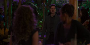 Movie still from “Sitting in Bars with Cake” (2023), directed by Trish Sie – A man standing in front of a group of people; Medium shot, Over the shoulder angle