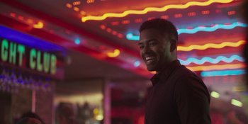 Movie still from “Sitting in Bars with Cake” (2023), directed by Trish Sie – A man smiles brightly in front of neon lights; Medium shot, Over the shoulder angle