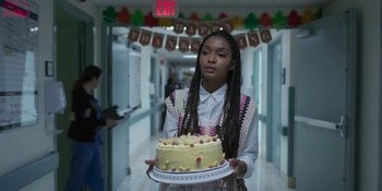 Movie still from “Sitting in Bars with Cake” (2023), directed by Trish Sie – A woman holding a cake on a plate in a hallway; Medium shot, Over the shoulder angle