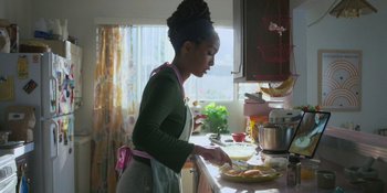 Movie still from “Sitting in Bars with Cake” (2023), directed by Trish Sie – A woman in an apron preparing food in a kitchen; Medium shot, Low angle