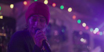 Movie still from “Sitting in Bars with Cake” (2023), directed by Trish Sie – A woman in a purple hat is singing into a microphone; Close Up shot, Over the shoulder angle