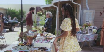 Movie still from “Sitting in Bars with Cake” (2023), directed by Trish Sie – A group of people standing around a table covered in food; Wide shot, Over the shoulder angle