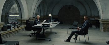 Movie still from “Skyfall” (2012), directed by Sam Mendes – Two people sitting at a table in an office; Wide shot, High angle