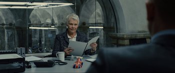 Movie still from “Skyfall” (2012), directed by Sam Mendes – An older woman sitting at a table holding a paper; Medium shot, Over the shoulder angle