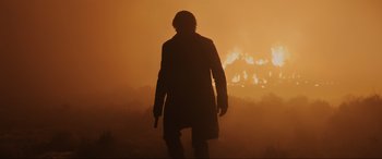 Movie still from “Skyfall” (2012), directed by Sam Mendes – A man in a trench coat standing in front of a fire; Wide shot, Over the shoulder angle