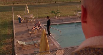 Movie still from “Bottle Rocket” (1996), directed by Wes Anderson – A man standing next to a swimming pool while a woman sits on a lawn chair next to the pool; Extreme Wide shot, High angle
