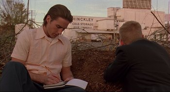 Movie still from “Bottle Rocket” (1996), directed by Wes Anderson – A man writing in a notebook while sitting next to another man; Medium shot, Low angle