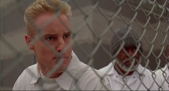 Movie still from “Bottle Rocket” (1996), directed by Wes Anderson – A man in a white shirt and a man in a gray hat and a fence; Close Up shot, Over the shoulder angle