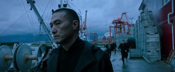 Movie still from “Snake Eyes” (2021), directed by Robert Schwentke – A man standing in front of a group of people in a harbor; Medium shot, Low angle