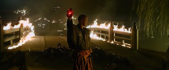 Movie still from “Snake Eyes” (2021), directed by Robert Schwentke – A man holding up a red object in front of a burning bridge; Medium shot, High angle