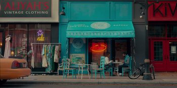 Movie still from “Sneakerella” (2022), directed by Elizabeth Allen Rosenbaum – Two people sitting at a table outside of a bakery; Extreme Wide shot, High angle