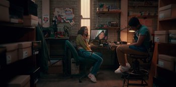 Movie still from “Sneakerella” (2022), directed by Elizabeth Allen Rosenbaum – A woman sitting on a chair next to a man in front of a television; Wide shot, High angle