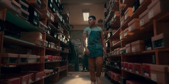 Movie still from “Sneakerella” (2022), directed by Elizabeth Allen Rosenbaum – A man wearing an apron walking through a room filled with boxes; Wide shot, Low angle