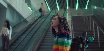Movie still from “Sneakerella” (2022), directed by Elizabeth Allen Rosenbaum – A woman in a colorful sweater standing in front of an escalator; Medium shot, Low angle