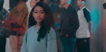 Movie still from “Sneakerella” (2022), directed by Elizabeth Allen Rosenbaum – A woman with long black curly hair standing in front of a group of people; Medium shot, Over the shoulder angle