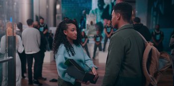 Movie still from “Sneakerella” (2022), directed by Elizabeth Allen Rosenbaum – A woman holding a box talking to a man in front of a group of people; Medium shot, Over the shoulder angle