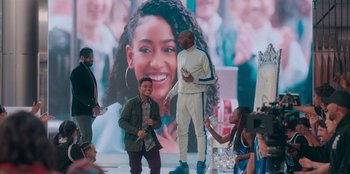 Movie still from “Sneakerella” (2022), directed by Elizabeth Allen Rosenbaum – Two men standing on a stage while a group of people look on; Wide shot, Over the shoulder angle